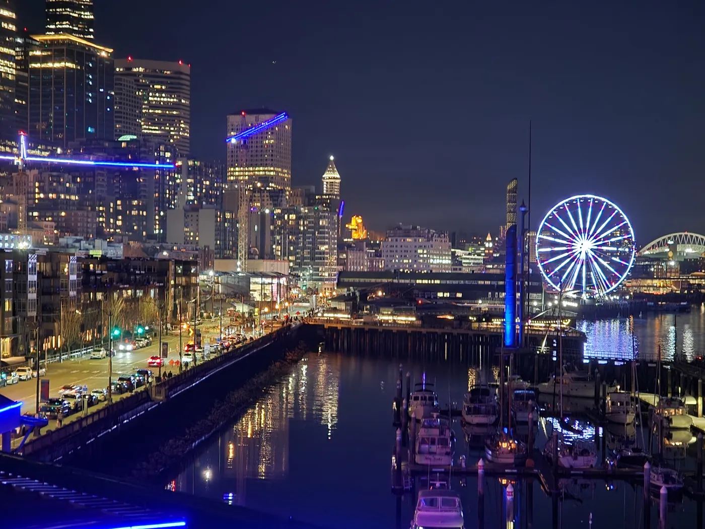 The Seattle waterfront at night — the Great Wheel lit up over Elliott Bay, boats in the marina, downtown towers glowing