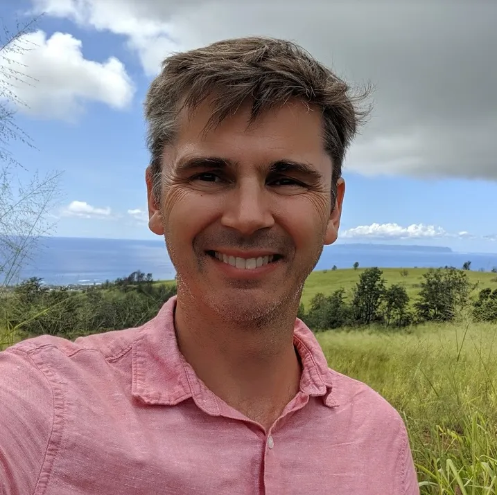 Jon Bostwick, smiling outdoors above green hills and the ocean in Maui