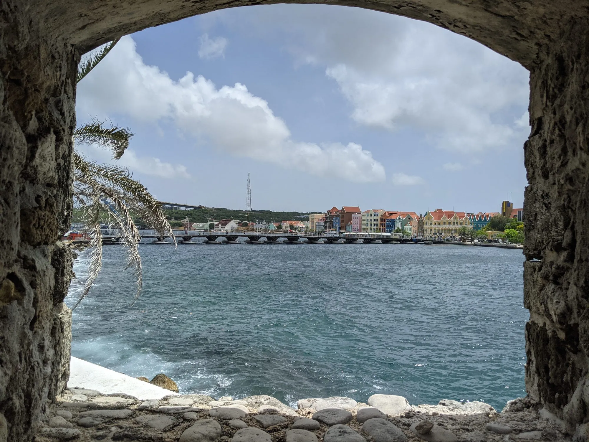 The pastel row of Willemstad, Curaçao — the Handelskade waterfront — framed through a stone fort window, a palm frond leaning in, turquoise sea beyond.